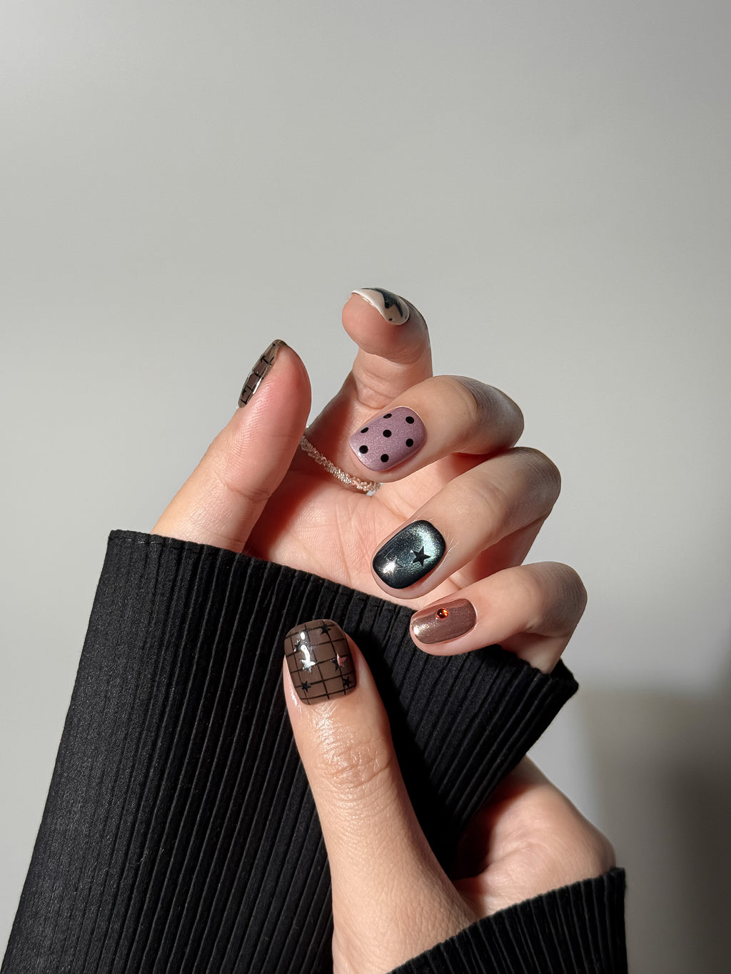 Close-up of a hand with a short square manicure wearing mismatched gel nails; the thumb features a dark brown grid pattern with silver stars, paired with black ribbed sleeve detail.