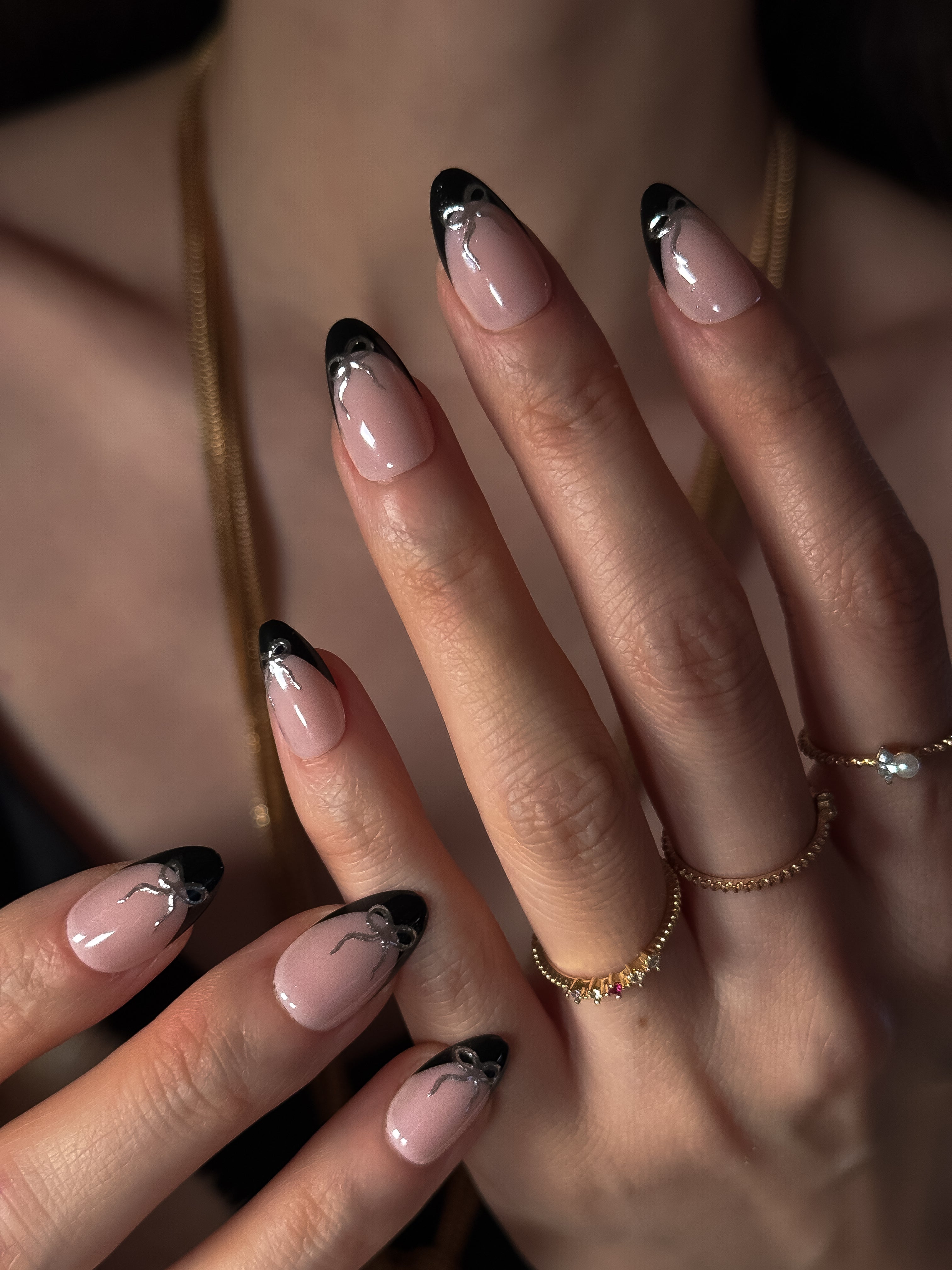 Close-up editorial shot of a hand wearing the Silver Ribbon Noir press-on nails, featuring black French tips and metallic silver bows, accessorized with gold rings and a gold necklace.