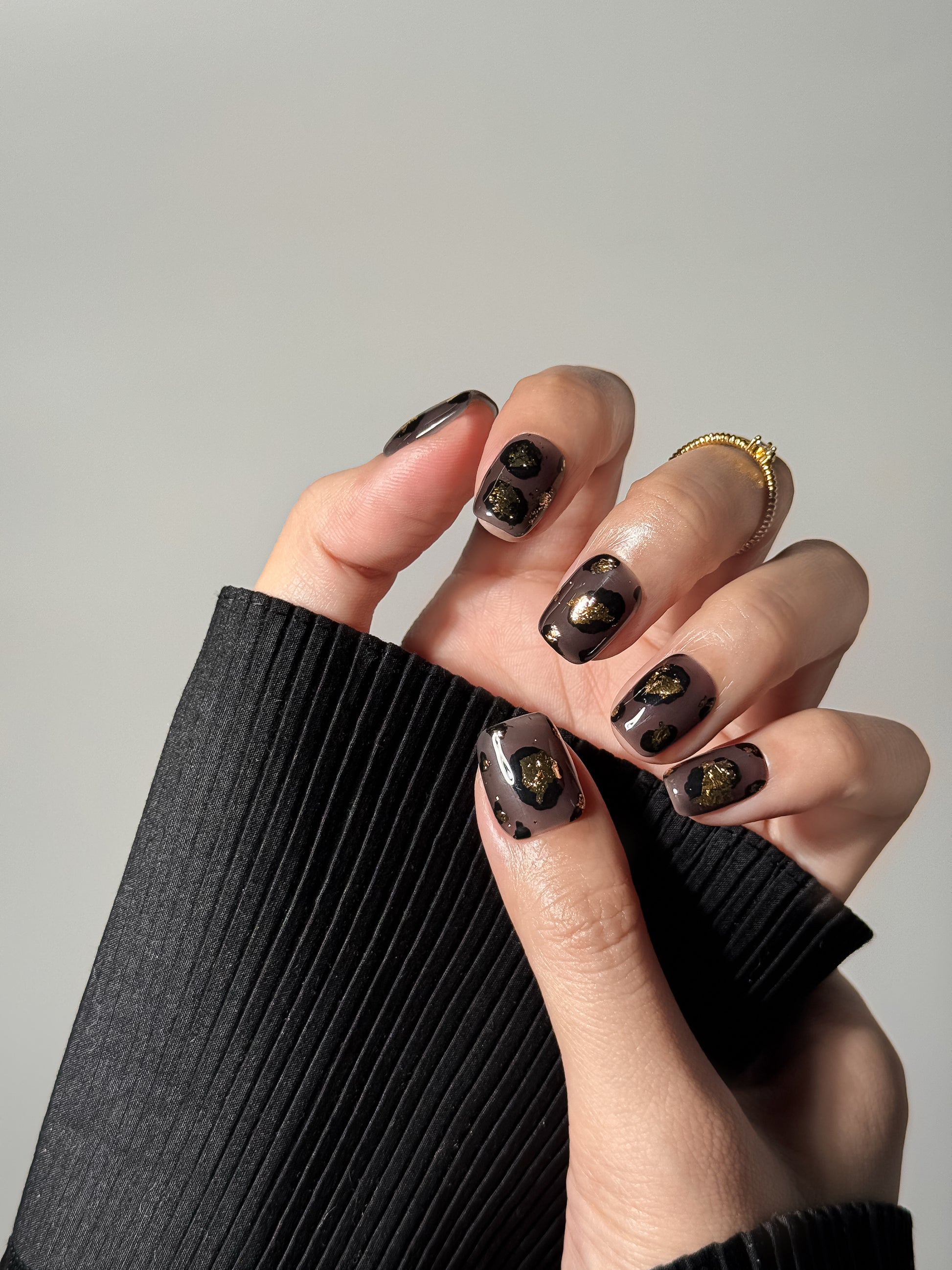 Hand wearing short square smoky grey nails with black leopard spots and metallic gold foil details, featuring a delicate gold ring.