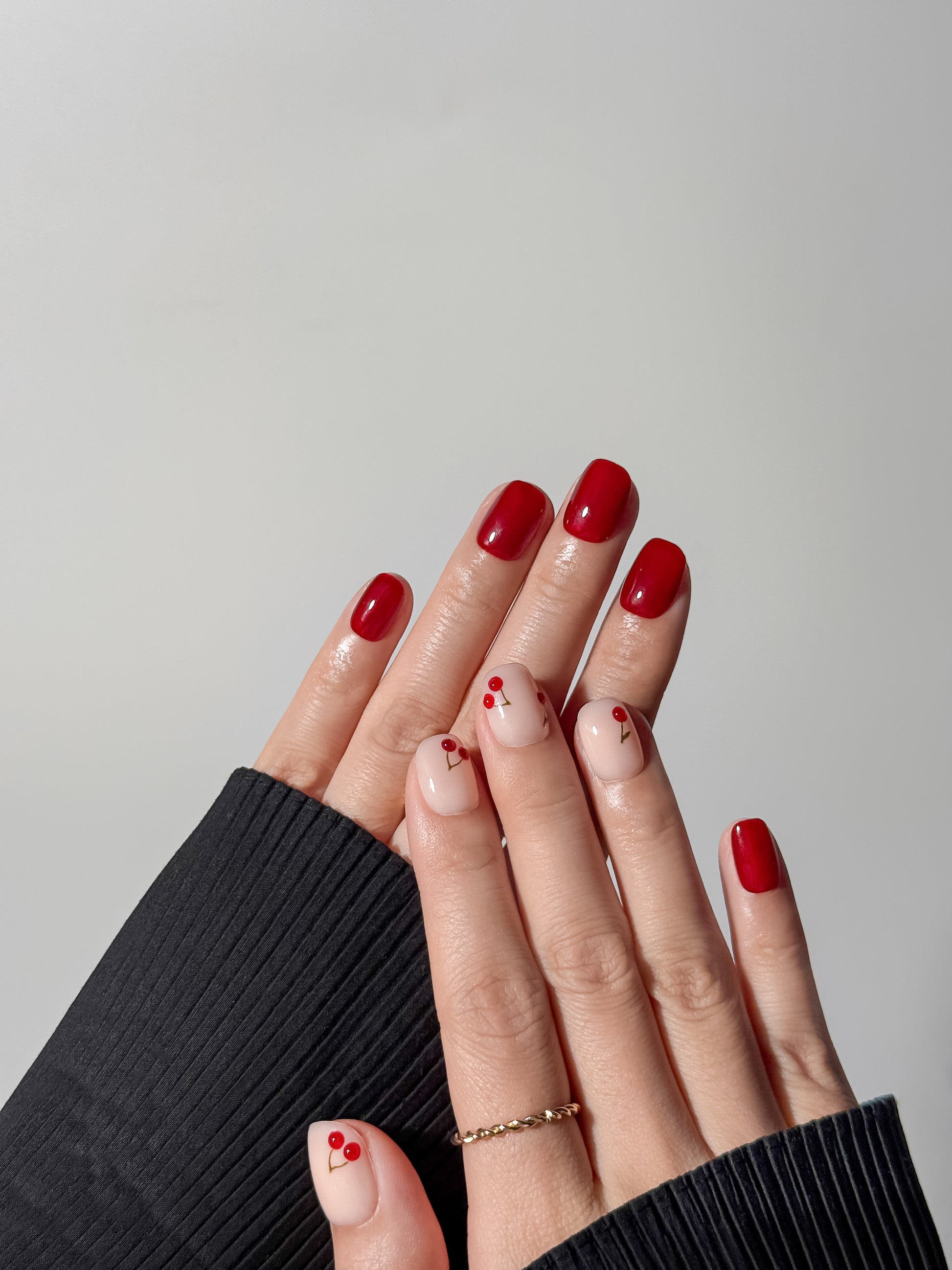 two hands intertwined showcasing a vibrant red manicure with dainty fruit-themed cherry nail art on the ring fingers and thumbs.