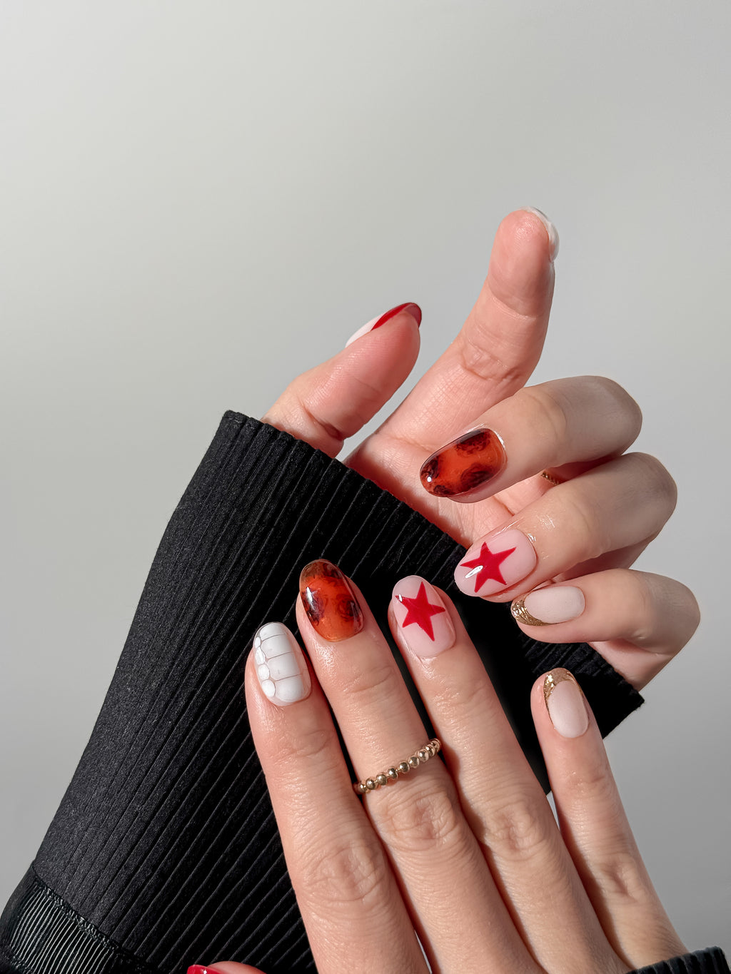 Two hands displayed against a black sweater showing a mismatched nail art aesthetic with vibrant red stars, amber tones, and gold glitter French tips on short oval nails. Reusable press-on nails from Justmee