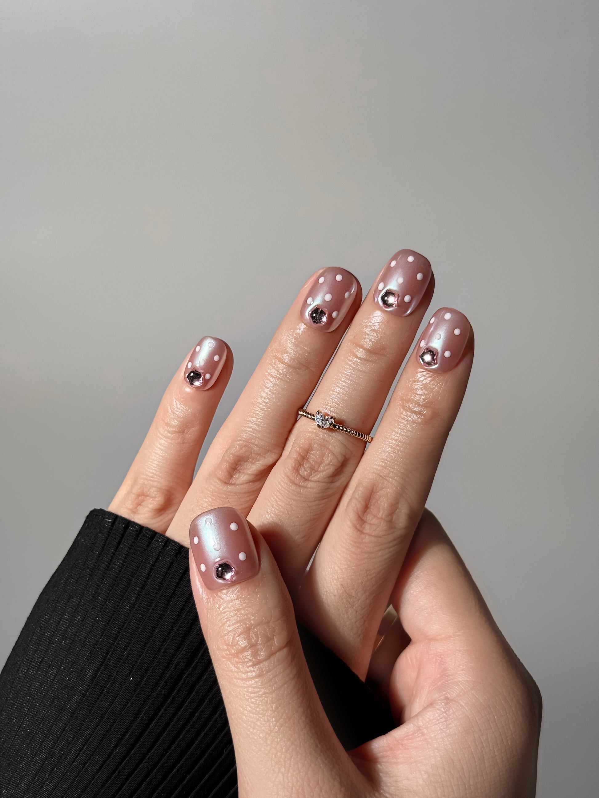 A hand wearing iridescent pink and white dotted nails with 3D pink crystals, styled with a gold heart ring and black sleeve.