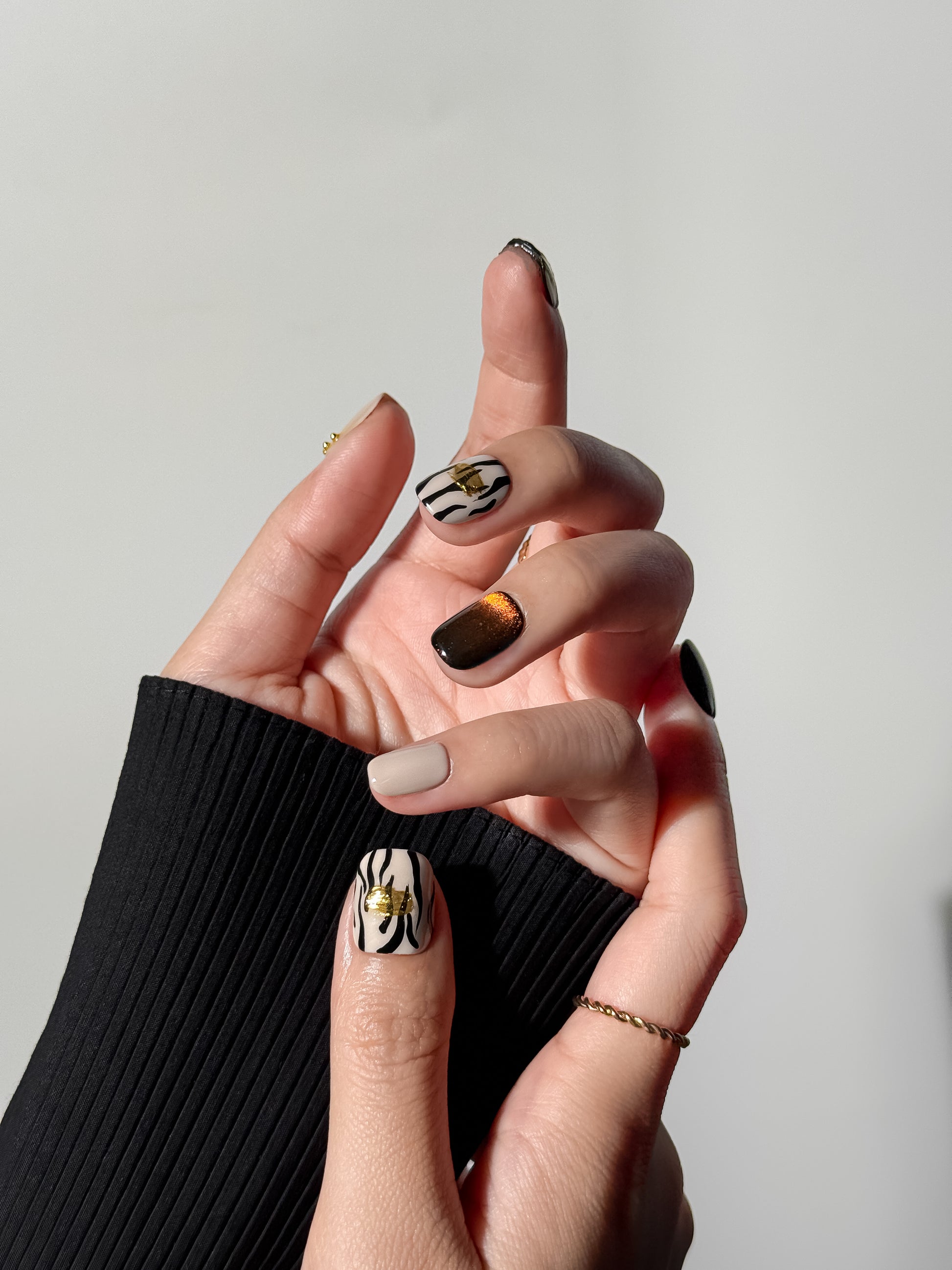 A close-up of a hand wearing short square nails; the thumb features zebra stripes with a 3D gold foil accent, and the middle finger shows a deep amber cat-eye magnetic gel effect against a black sleeve.
