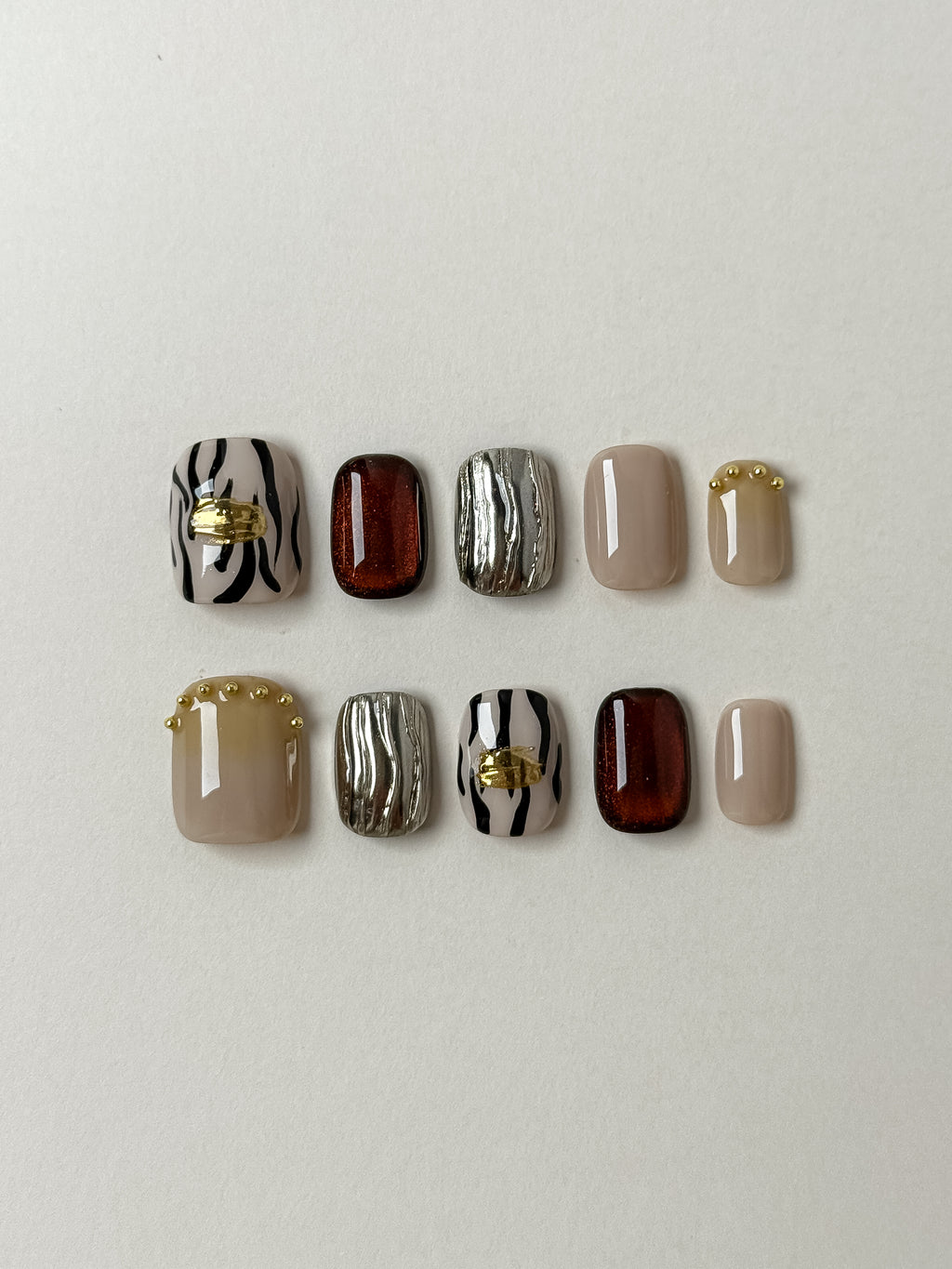 A flat lay set of ten handmade press-on nails featuring a mix of black and white zebra stripes with gold leaf, amber cat-eye magnetic polish, textured 3D silver chrome, and nude beige with gold micro-beads.