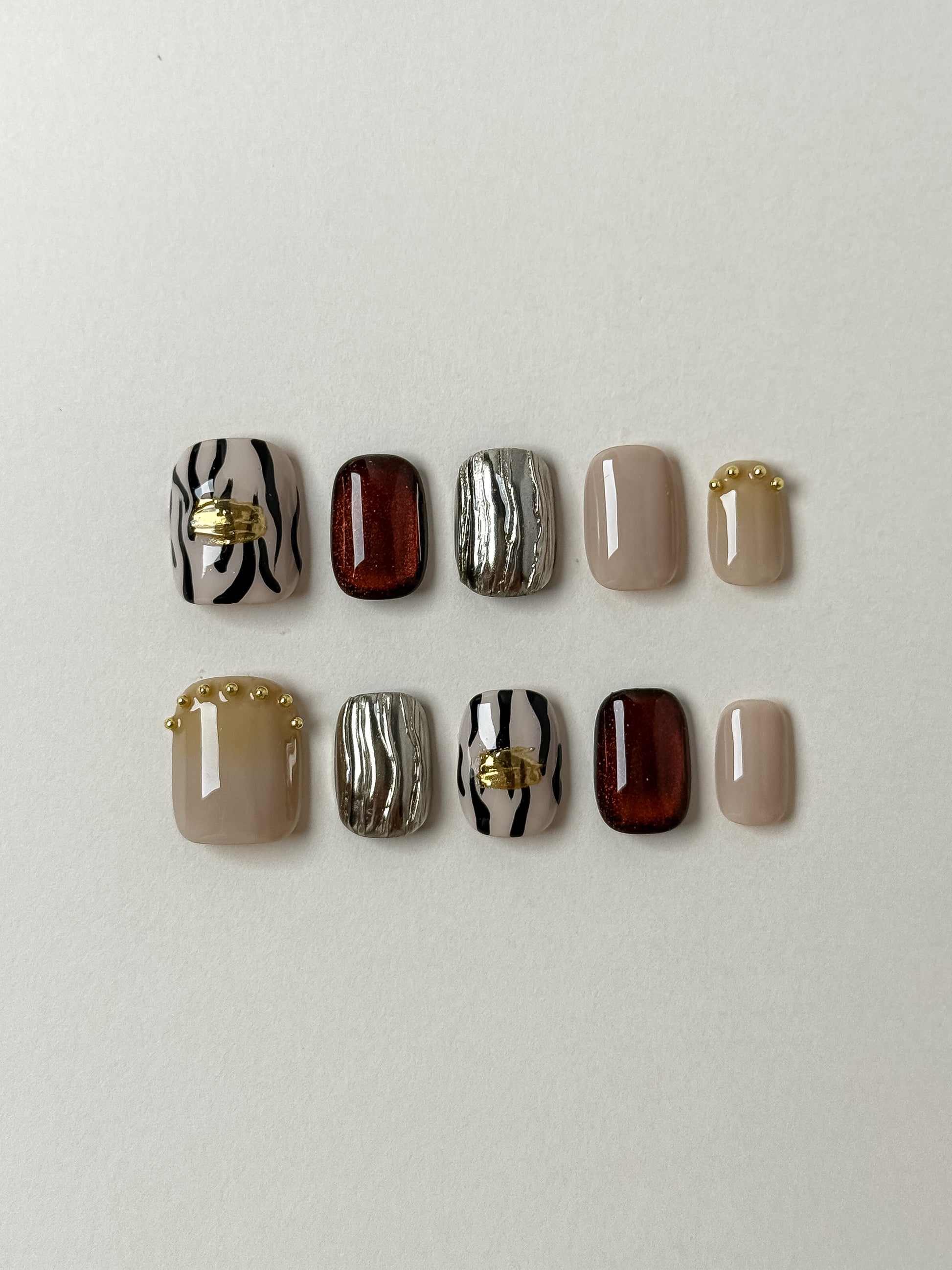 A flat lay set of ten handmade press-on nails featuring a mix of black and white zebra stripes with gold leaf, amber cat-eye magnetic polish, textured 3D silver chrome, and nude beige with gold micro-beads.
